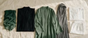Flat lay of Muslim women's garments on cream linen: green chiffon hijab, black abaya, sage open abaya, grey hooded khimar and white two-piece prayer set